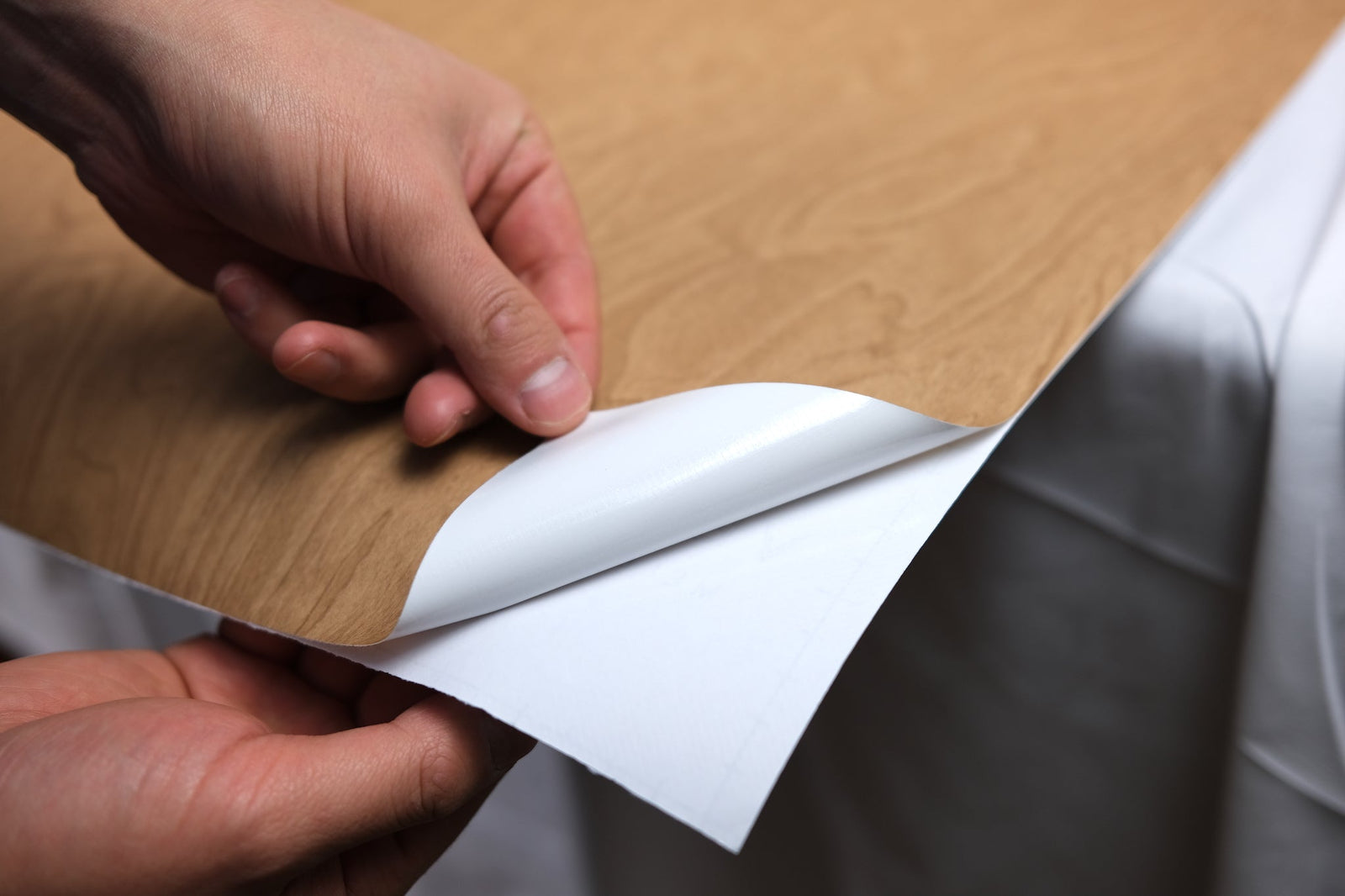 Hands peeling the white backing paper off a light oak wrap sheet over a clean surface