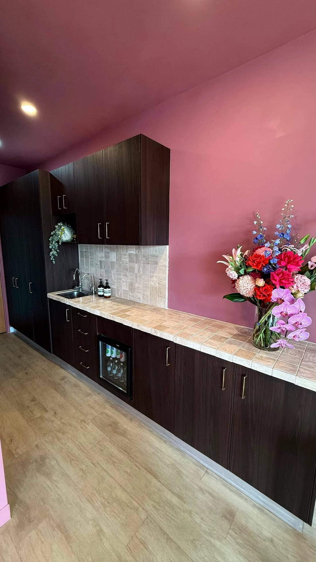 Kitchenette cabinet doors wrapped in Chocolate Walnut vinyl against a pink wall, with a stone splashback and fresh flowers