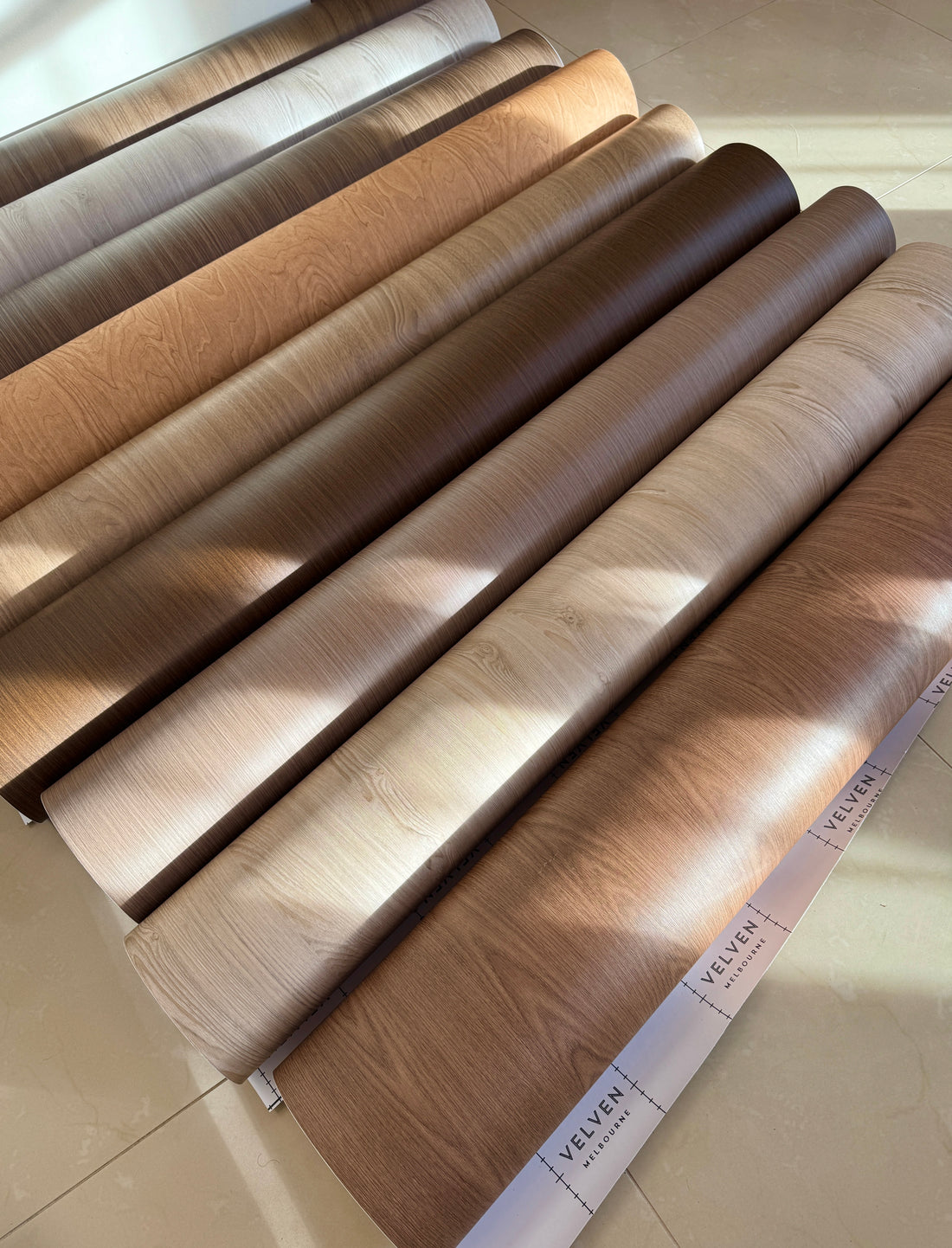 Woodgrain vinyl wrap rolls in oak and walnut tones arranged in natural light