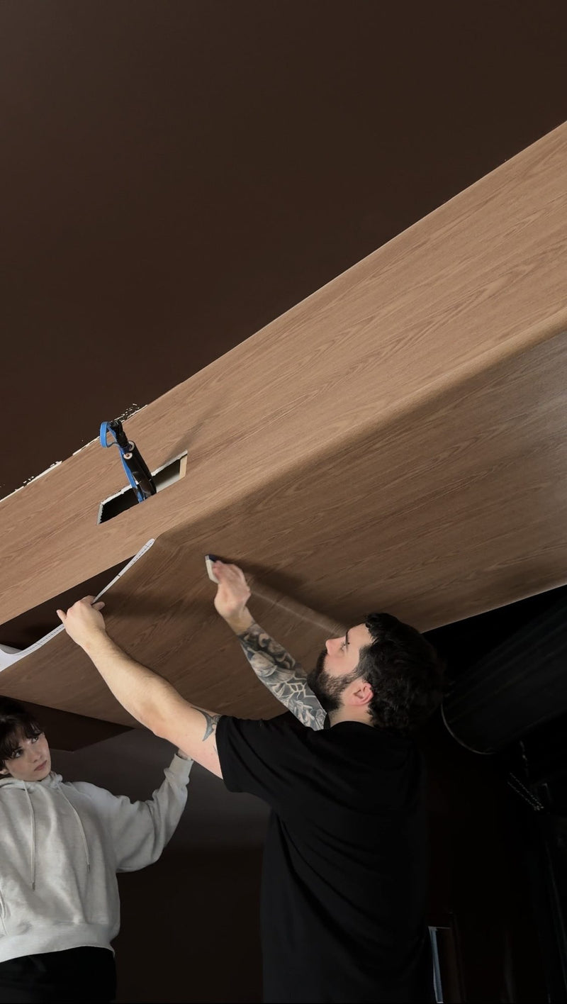 Two installers applying a light wood grain vinyl panel to a dark ceiling