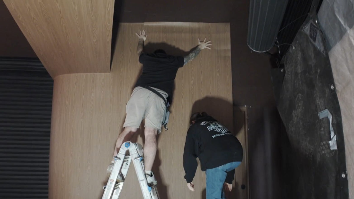 Installers on a ladder applying Maison Walnut wood grain wrap to a tall feature wall at The Feel Good Hood